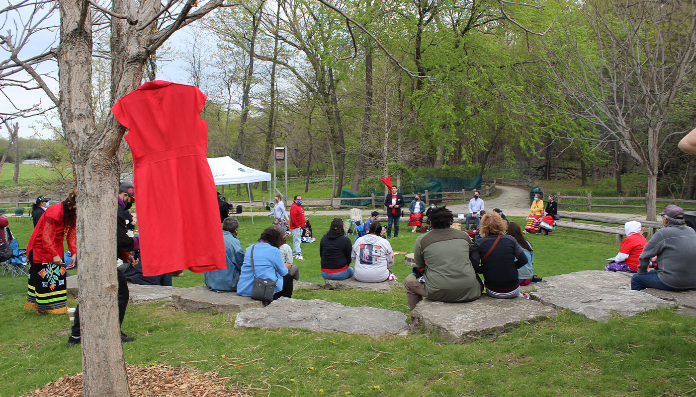 a group of people gathered around a speaker, a red dress hangs in the foreground from a tree