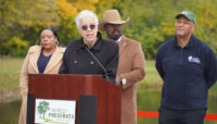 A group of people presenting at a podium in front of a ribbon, adjacent to a body of water.
