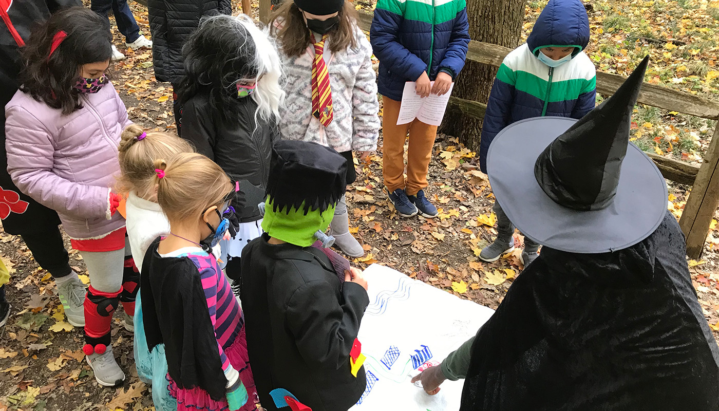 A group of children and an adult, all in Halloween costumes, gather around a map.