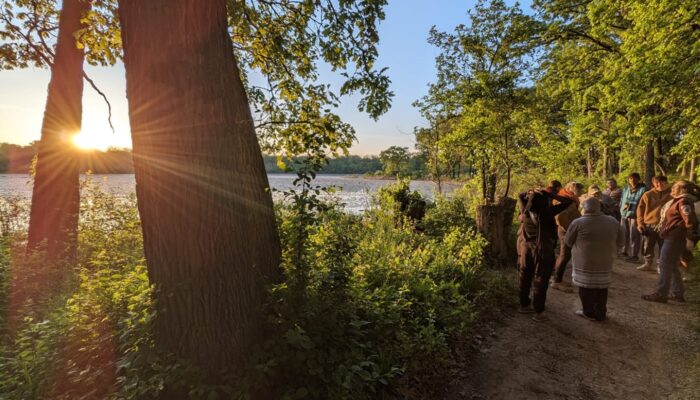 A group of people standing near a body of water during sunrise along a forested trail.