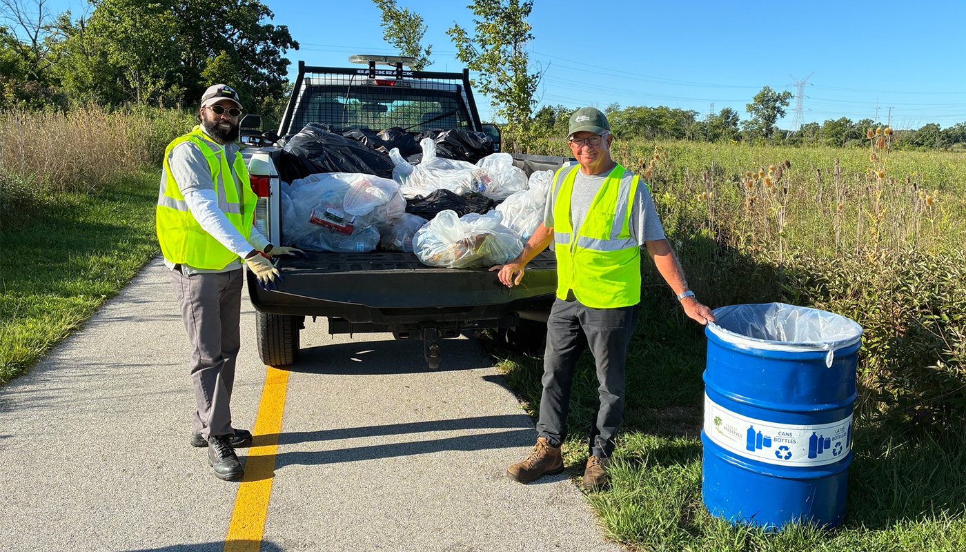 two people wearing safety vests standing in front of the bed of a truck filled with garbage bags