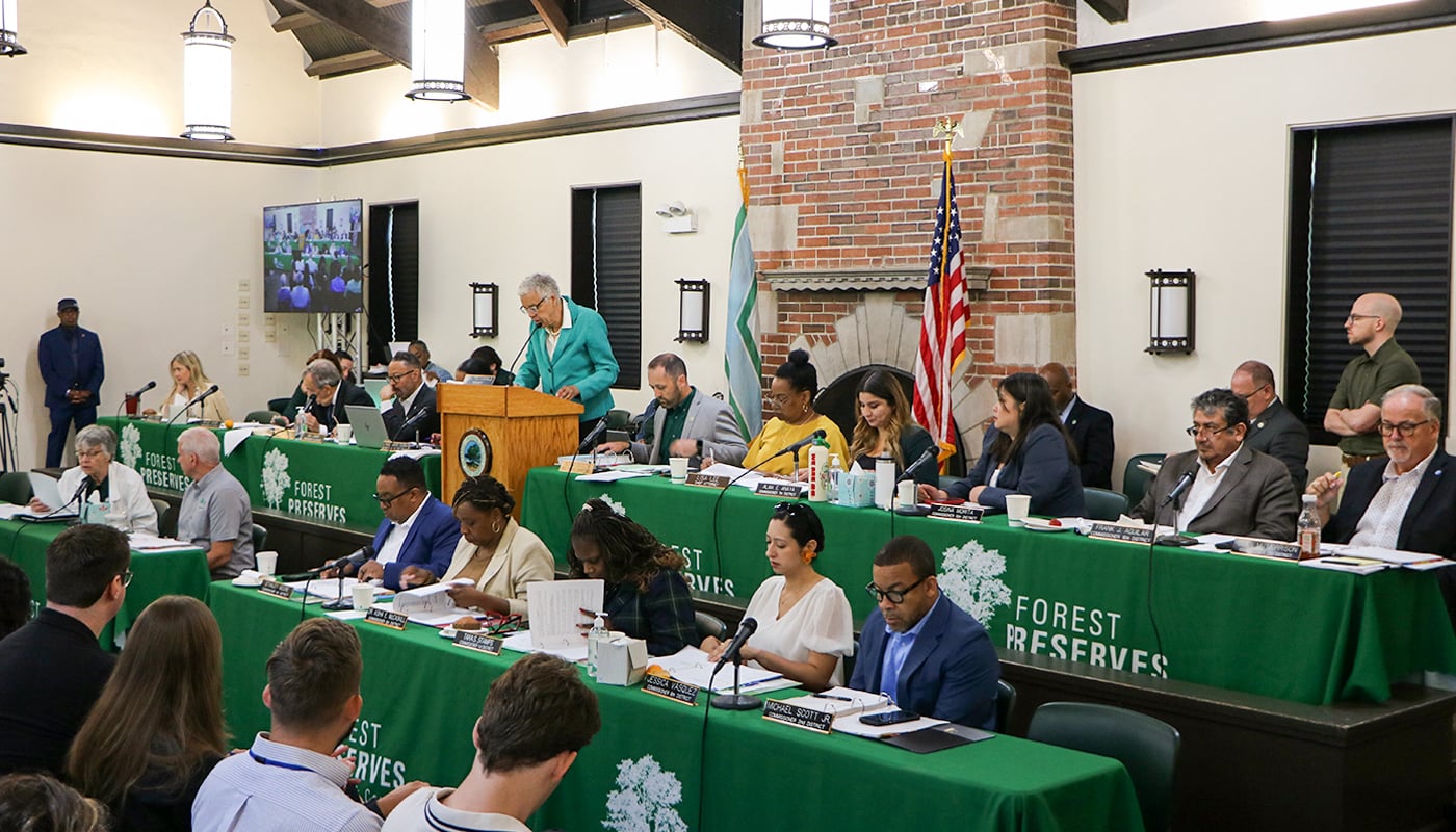 Cook County Board President Toni Preckwinkle speaks at a podium during a Board of Commissioners meeting, with Forest Preserves Superintendent Adam Bianchi and commissioners seated nearby.