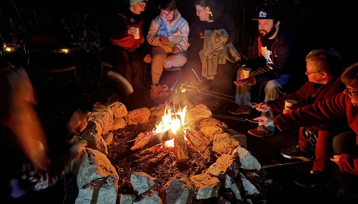 A group of people surrounding a campfire and roasting marshmallows.